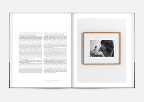 An overhead shot of an open book laid flat. The left page contains two columns of black text. The right page is mostly blank, featuring a single, wood-framed, black-and-white photograph. The photograph shows a young person with dark hair looking down at a miniature, topless woman standing in their cupped hands.