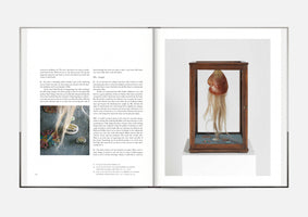 A photograph of an open art book laid flat. The left page contains black text with a small inset image. The right page features a full-bleed photograph of a sculpture in a wooden and glass vitrine. The artwork is a fleshy, biomorphic object with long blonde hair hanging from it.