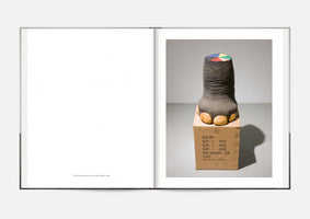 An open art book shows a photograph on the right-hand page. In a grey studio, a sculpture of a severed elephant's leg stands on a cardboard shipping box marked with MADE IN CHINA. The leg has realistic wrinkled grey skin and yellowed toenails, but the flat, severed top is painted with a colorful pie chart. The left page is blank except for a caption at the bottom.