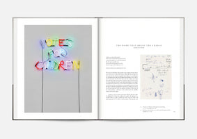 An open book viewed from above. The left page features a photograph of a neon sign that reads VOTES FOR CHILDREN in multi-colored, child-like handwriting. The right page is titled THE HAND THAT ROCKS THE CRADLE and contains an essay alongside a reproduction of a handwritten sketch.