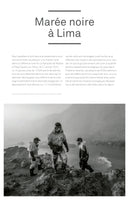 A black and white photo shows three workers in oil-stained protective suits and respirators walking along a rocky, slick shoreline. They are part of a cleanup crew responding to an oil spill, with the sea visible in the hazy background.