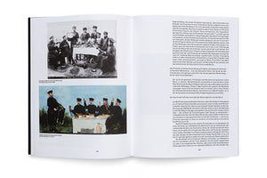 A close-up photograph of a two-page spread in an open book. The left page displays a vintage black-and-white photograph of men at an outdoor feast above a colorful, naive-style painting of a similar scene. The right page consists of columns of German text.