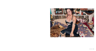 A young woman with dark curly hair and tattoos sits on her bed in a cluttered room, wearing a black satin slip with lace trim. She looks down towards a tabby cat that is partially hidden by the leopard print and floral patterned blankets and is looking at the camera. The wall behind her is decorated with photos and personal items.