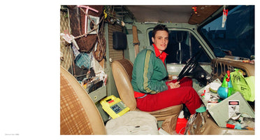 A person with short dark hair sits in the driver's seat of a heavily cluttered van, looking directly at the camera with a slight smile. They are wearing a green jacket with yellow stripes over a red collared shirt and red pants. The van's interior is packed to the ceiling with personal belongings, tools, and various items, filling the passenger seat, dashboard, and the space behind them.