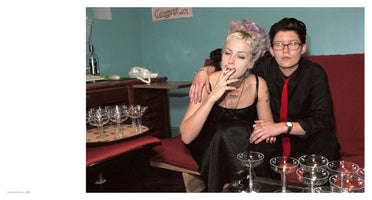 A color photo from 1998 titled Amita and Sunny. A woman with short blonde and purple hair in a black dress smokes a cigarette with her eyes closed while sitting on a red couch. A person next to her with short dark hair, glasses, and a red tie has their arm around her and looks directly at the camera. The room has teal walls and is filled with empty champagne coupe glasses on tables around them.