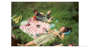 A high-angle, sunlit photograph of several people having a picnic in a field of long green grass. In the foreground, a woman in a green dress lies on her back with her eyes closed. Behind her, another person with short hair sits up on a red and white checkered blanket, looking at the camera. A small, dark dog sits near the blanket next to picnic supplies. The top of a third person's head is visible at the bottom of the frame.