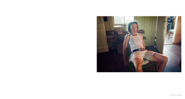 An interior, full-length color photograph of a person with short, dark, curly hair sitting in a relaxed posture on a green wooden chair. They wear a white tank top, light shorts, and a dark belt, and they look directly at the camera. The room is rustic and lit by natural light from a window in the background.
