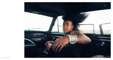 A film photograph taken from inside a vintage car, showing a Black person in the passenger seat looking back over their shoulder. Their large afro is blown sideways by the wind. They have tattoos on their arm and wear a thick, coiled silver bracelet. The bright landscape outside is visible through the open windows.