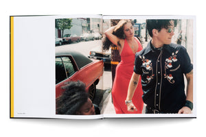 A color photograph from 1996, displayed in an open book, shows a candid street scene. A man in a black, floral-embroidered shirt and sunglasses stands in profile next to a woman in a vibrant red dress who is adjusting her hair. In the foreground, the top of another person's head with curly hair is visible beside a vintage red car.