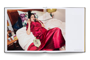 A full-page color photograph in an open book showing a woman with dark hair and red lipstick wearing a shiny red satin robe. She is lying in bed, looking up while talking on a green landline phone and holding a cigarette. A leaf-shaped ashtray rests on her lap, and her bedside table is cluttered.