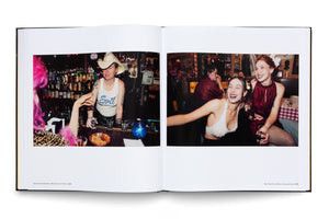 An open book with a black cover displays two color photographs from the late 1990s. The photo on the left shows a bartender with tattoos and a cowboy hat serving drinks. The photo on the right shows two young women laughing and smiling while seated at a table in a dimly lit restaurant.