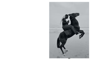A black and white photograph of a shirtless Black man with white angel wings, riding a dark horse that is rearing up on a beach next to the ocean.