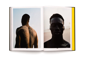 An open black photobook on a white surface. The left page shows a photo of a muscular Black man from behind, his back covered in sand, silhouetted against a light blue sky. The right page shows a silhouetted close-up portrait of a Black man's face against a pale sky.