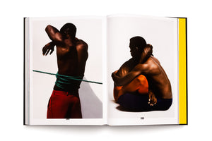 An open book on a white background showing a two-page spread of color photographs. On the left, a muscular Black man in red trousers is seen from behind, his torso wrapped in a green rope. On the right, two muscular Black men, one in orange and one in blue pants, are seated in an intimate embrace.