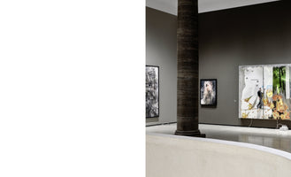 An interior shot of a modern art gallery with grey walls and a polished floor. A large, dark, textured column stands in the center, with various artworks hanging on the walls behind it, including a large, illuminated multi-panel piece and a smaller portrait. In the foreground is the edge of a curved white bench or wall.