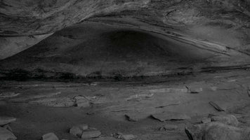 A wide, black-and-white photograph of the interior of a large, shallow rock cave. The ceiling and back wall are composed of sweeping, layered stone, with the back receding into a deep, dark alcove. The uneven floor is scattered with rocks and features a low, rough wall made of rectangular stone blocks.