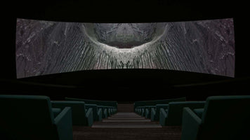 A low-angle view from the back of a dark, empty movie theater, looking down a central aisle between rows of teal seats. A massive, curved screen displays a stark, symmetrical, black-and-white image of a rocky volcanic crater, creating an abstract and imposing shape that resembles a giant eye or a face.