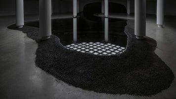 A minimalist art installation in a dimly lit room with a concrete floor and several white pillars. A meandering pool of dark, reflective liquid, bordered by black gravel, flows through the space. A grid of white squares glows from beneath the surface of the liquid.