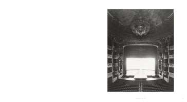A high-angle, black-and-white photograph of the lavish interior of an empty opera house, the Palais Garnier in Paris. The view from an upper balcony looks down on rows of seats, ornate boxes lining the walls, a grand chandelier, and a stage that is completely illuminated in white light.