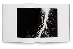 An open book on a white surface, displaying a striking black-and-white photograph of a powerful lightning bolt against a pitch-black background. The bright white bolt and its many fine, branching tendrils fill the right-hand page, while the left page is mostly blank white space.