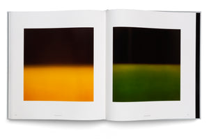 An overhead shot of an open art book on a white surface. The two-page spread displays two minimalist, abstract photographs. The image on the left features a dark top half blending into a vibrant orange bottom half. The image on the right has a similar composition with a dark top half blending into a deep green bottom half.