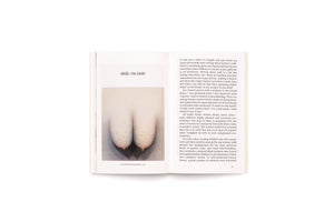 A photograph of an open book on a white surface. The left page features the title Milk: On Debt and a stylized painting of two breasts by Loie Hollowell. The right page contains an essay about mother's milk.