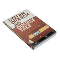 A paperback book titled Stefan Draschan An Double Take, lying diagonally on a green background. The cover image shows the rear of a brown and tan vintage camper trailer parked in front of a red brick wall. The author's name and title are printed in large, brown, sans-serif letters, appearing as if they are part of a sign on the building.