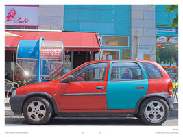 A street-level, side-view photograph of a two-tone car parked on a Berlin street. The front half of the small hatchback is red, matching the red awning of a café in the background, while the back half is a bright turquoise, matching the facade of the building next to the café. An older man is visible sitting in the driver's seat.
