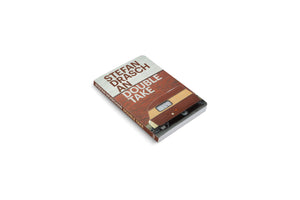A high-angle shot of the book An Double Take by Stefan Draschan on a plain white background. The book cover features a photo of a vintage brown and tan camper parked in front of a red brick wall.