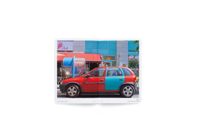 A photograph in an open book shows a street scene in Berlin. A small car, painted red on its front half and turquoise on its rear half, is parked in front of two storefronts. Behind the car, a café with a red awning matches the car's front, and an adjacent building with a turquoise facade matches the car's rear.