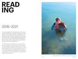 A book page with a two-column layout. On the left, against a white background, is the word READING in large black letters, followed by the dates 2016-2021 and descriptive text. On the right is a color photograph of a man standing chest-deep in calm, blue-green water, fully clothed and wearing glasses, as he reads a pink book titled das, kapital.