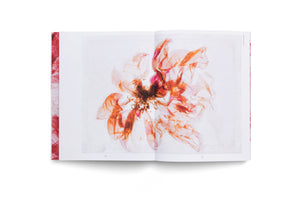 An open book with a red textured spine lies flat on a white surface. The pages display a large, ethereal image of a flower with translucent, delicate petals in shades of orange, red, and pink, with a dark, fibrous center.