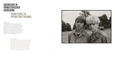 A two-page book spread. The left page has text in German and English with the title YOUNG PEOPLE IN PRE-MILITARY TRAINING. The right page features a black-and-white photograph of two serious-looking young men in uniform shirts. The man on the left has dark, straight hair and a mustache, while the man on the right has a full head of curly, light-blonde hair. They are outdoors in a field with trees in the background.