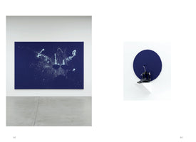 A two-page spread from an art book. On the left, a large, dark blue rectangular painting hangs on a white gallery wall. The painting is covered in white splatters that form the abstract shape of a drum kit in motion. On the right, a smaller installation shows a drum pedal connected to a blue paint roller, positioned in front of a blue painted circle on the wall, revealing the artistic process.