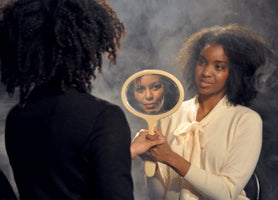 A studio portrait of two Black women with voluminous natural hair against a smoky grey background. The woman on the left is seen from behind, while the woman on the right, in a cream blouse, holds an oval hand mirror with her. The reflection in the mirror shows a woman's face staring directly forward.