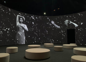 A dark, round room with an immersive video installation. A large, curved screen displays a black-and-white projection of two people, one posing in a dress and the other waving, against a dark background with floating white specks. Several low, round, light-colored stools are arranged on the floor.