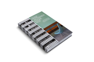A thick hardcover book titled Arne Jacobsen Room 606 by Michael Sheridan, laid diagonally on a white surface. The book's cover is split: the spine and left side have a graphic black and white tile pattern, while the right side is green and features a photograph of a modern, light blue chair in a room.