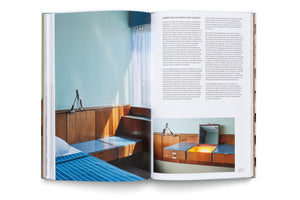 An overhead view of an open book showing a two-page spread. The left page features a large photo of a mid-century modern room with a bed, a blue striped blanket, and built-in wooden furniture against a light blue wall. The right page contains German text and a smaller photo of a wooden vanity with a built-in light. The page number 207 is visible at the bottom.