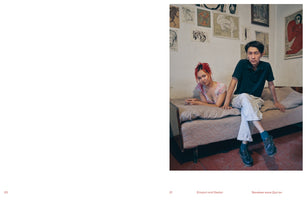 A two-page spread from a publication. The left page is blank. The right page features a full-bleed photograph of a young woman with vibrant red hair and a young man sitting together on a simple bed in a sparsely furnished room. The wall behind them is covered with numerous sketches and drawings. Both individuals look directly at the camera with neutral expressions.