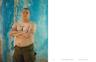 A shirtless person with a buzz cut and numerous tattoos stands with their arms crossed in front of a textured, bright blue wall. They wear green cargo pants and look directly at the camera with a serious expression. Notable tattoos include a large cross on the chest, rifles on the forearms, and a spiderweb on an elbow. The image is on the left page of a book spread.