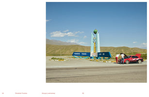 A color photograph of a rural roadside on a sunny day. Two people are looking under the open hood of a red sports car parked next to a large, blue monument with Cyrillic text. In the background, rolling green hills and mountains rise under a clear blue sky.
