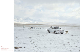 A silver sedan and a dog stand in a vast, snow-covered plain under a cloudy sky. In the background, a range of tall, snow-capped mountains stretches across the horizon. The image is on a book page with the caption, Song Kul in Summer—A Snowy Surprise.