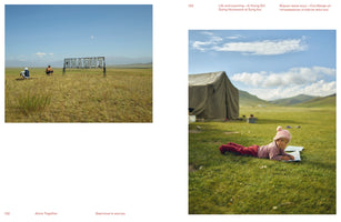 A two-page book spread with two photographs. On the left, a wide shot shows two people sitting in a vast, dry grassy field with a large metal sign and mountains in the distance. On the right, a young girl in a pink hat lies on lush green grass, looking up from a notebook, with a large canvas tent behind her.