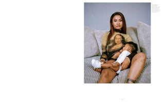 A woman wearing a sheer, long-sleeved shirt with the Mona Lisa printed on it sits on a grey corduroy couch, looking directly at the camera. She is breastfeeding a baby in a white onesie and a single white sneaker who rests in her lap. The woman has long brown hair and visible tattoos on her arms and legs.