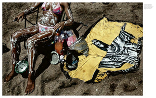 A high-angle photograph of a person with dark skin sitting in a folding chair on a dark sandy beach. They wear a pink patterned bikini and their body is liberally covered in a thick white foamy substance. Next to the chair on the sand is a crumpled yellow towel with a black-and-white graphic of the Christ the Redeemer statue.