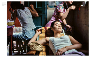 A candid color photograph of three young Asian women in a cramped, dimly lit room. In the foreground, a woman in a white tank top rests with her eyes closed in a wooden chair. Behind her, another woman in a pink patterned top reaches for an electrical outlet, while a third woman with her back to the camera sits at a small table.