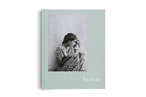 A light greenish-blue hardcover book titled human in white lowercase letters. The cover features a black and white photograph of a young person in a floral dress, sitting against a cracked wall while looking at their reflection in a small, hand-held mirror.