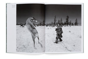 A black-and-white photograph spread across two pages of an open book. In a snowy landscape, a person in a heavy winter coat uses a rope and a long pole to handle a reindeer that is leaping high into the air. A large herd of reindeer and a line of sparse trees are visible in the background under a dark sky.
