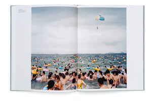 An open photobook displays a two-page spread showing an extremely crowded sea under an overcast sky. Hundreds of people are packed into the water, many using colorful inflatable rings, while a single person parasails high above the crowd.