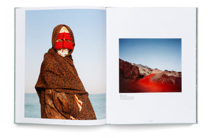 An open photobook spread on a white background. The left page displays a portrait of a person wearing a patterned brown cloak and a vibrant red mask that covers their face, standing by the sea. The right page shows a landscape photo of a bright red road snaking through rocky hills, with descriptive text underneath.